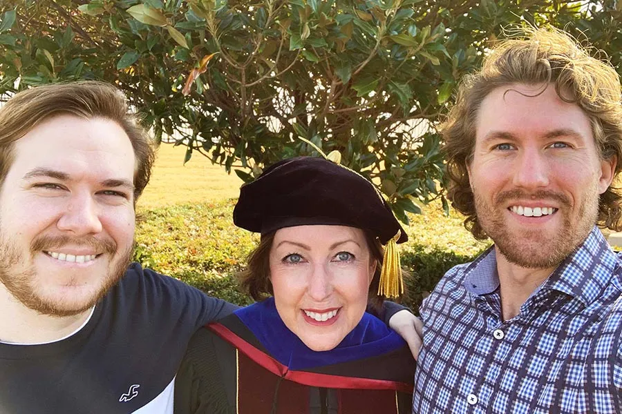 a OT PhD graduate dressed in graduation regalia stands between two sons in front of a tree