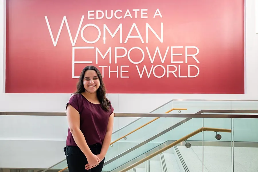 Student Regent Alyssa Flores stands in front of a staircase with Educate a Woman Empower the World mural