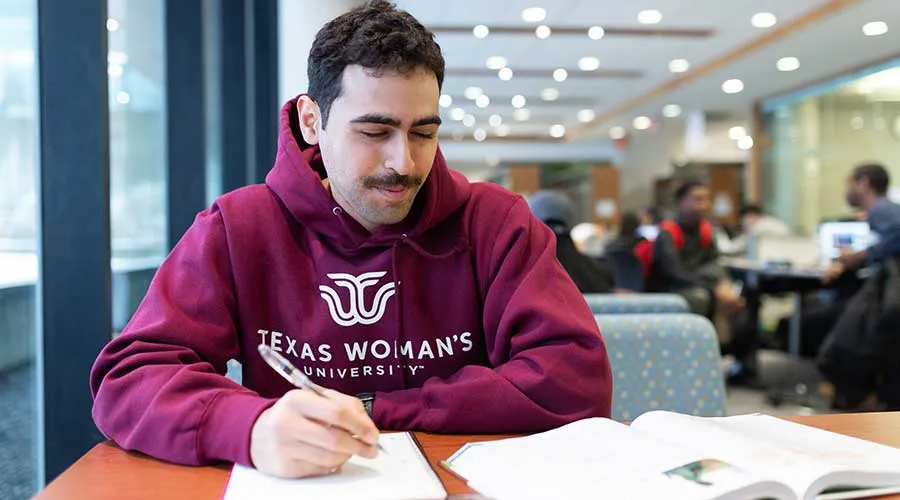 A male TWU student in a TWU hoody studies a textbook in a library setting.