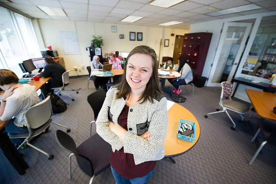 A young woman smiles as tables of students in work groups collaborate in the background