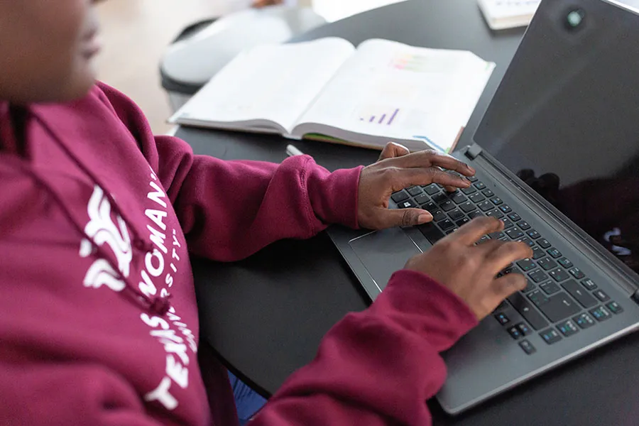 A student wearing a TWU sweatshirt works on a laptop with an open book nearby