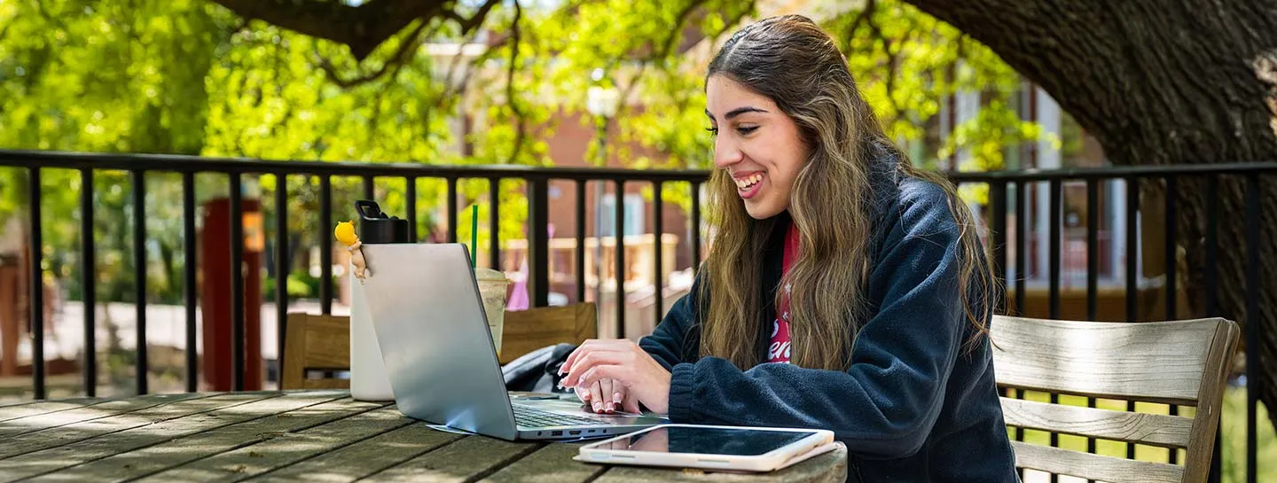A smiling TWU student sits outside by the library fountain while working on a laptop