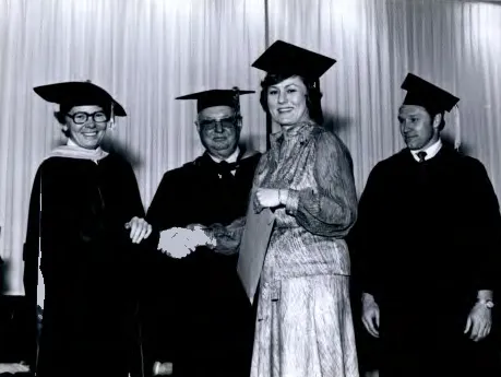 A group of faculty in regalia shake hands with a student, formal, black and white photo