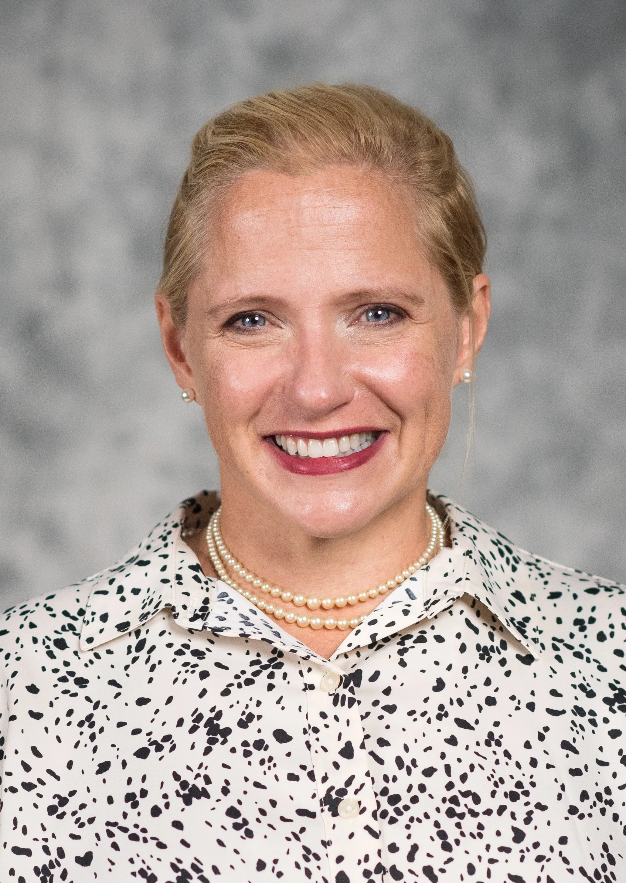 woman with pulled back blonde hair smiles directly at camera in front of grey background