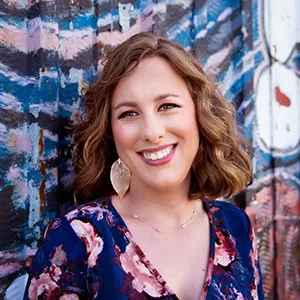 Blonde woman with shoulder length hair and a floral dress smiles at the camera in front of a colorful mural