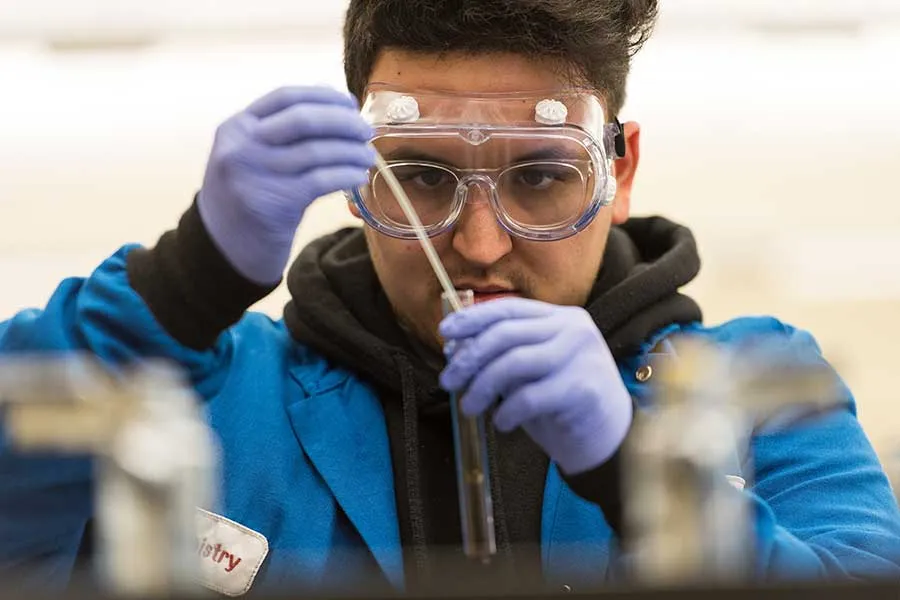 Chemistry student in lab coat, gloves, goggles pipetting into a vial