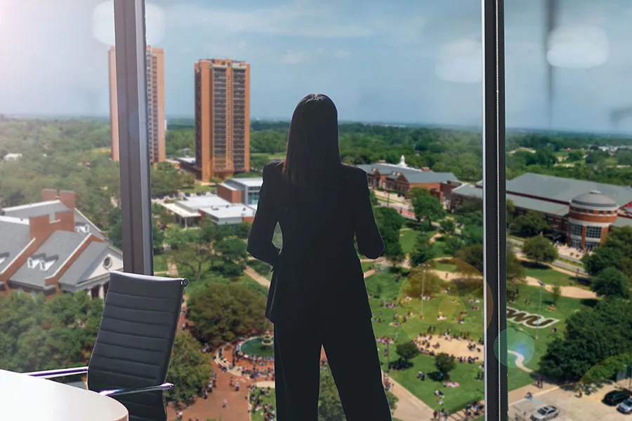 The silhouette of a woman stands at a full-length window overlooking the TWU Denton campus far below