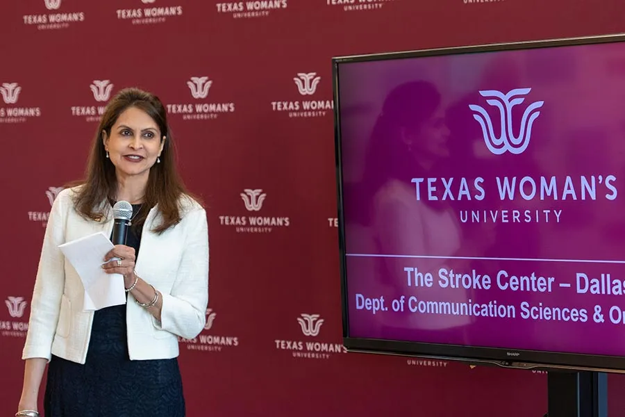 Dr. Mehta holds a microphone in front of a TWU backdrop next to a monitor