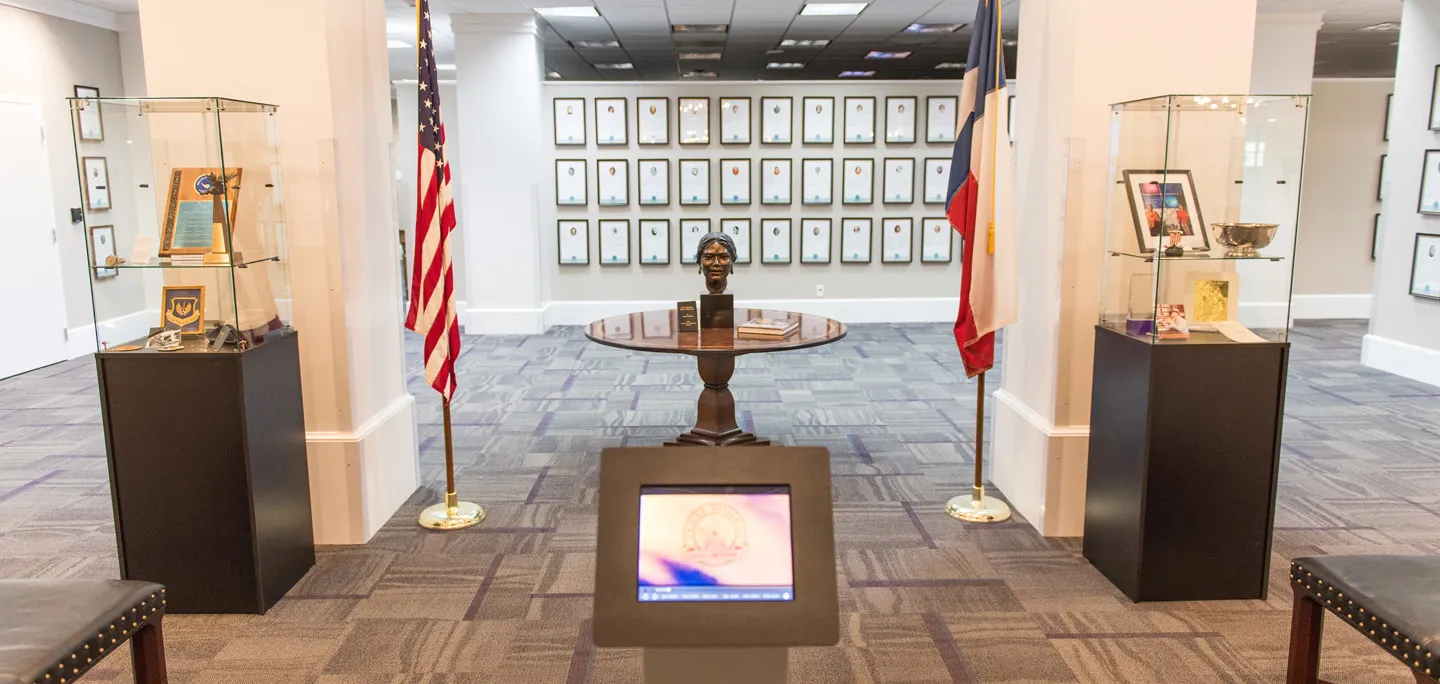The Texas Women's Hall of Fame, with a statue sitting on a table with flags on either side. Plaques line the wall in the background.