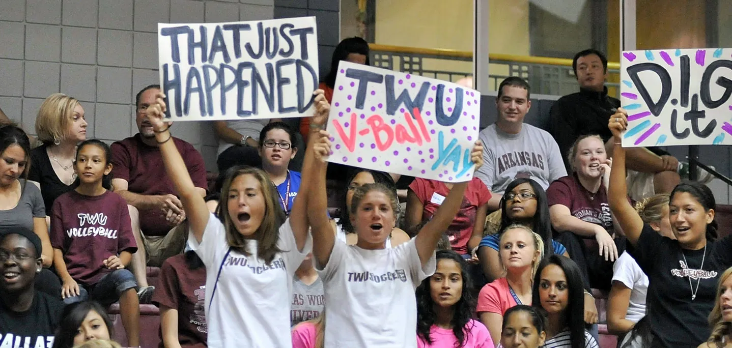 A group of students in bleachers holding up signs and cheering for a volleyball game