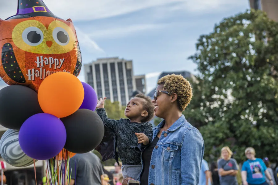 A mother holding her child while he plays with a balloon that says 'Happy Halloween' on it.