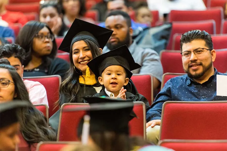 A child sits on the lap of a TWU student dressed in academic regalia in a crowd.
