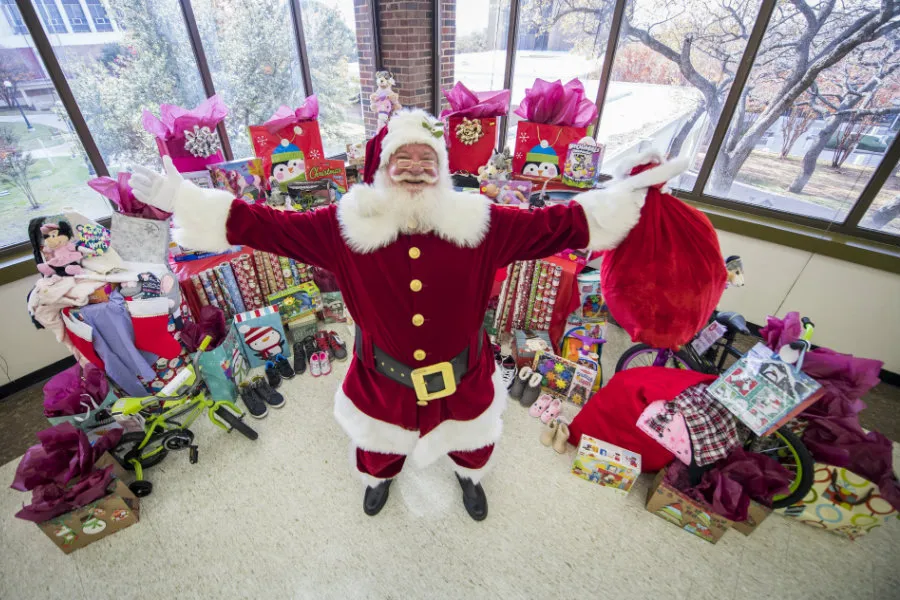 Santa standing with his arms out in front of a pile of toys and presents on TWU's Denton campus.