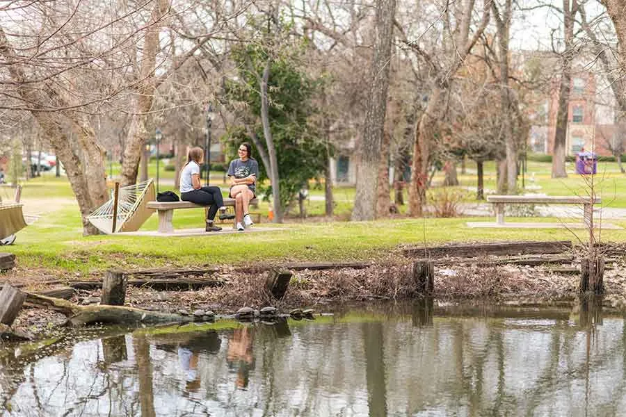 Two TWU students relax and sit on a bench outdoors on the Denton campus.