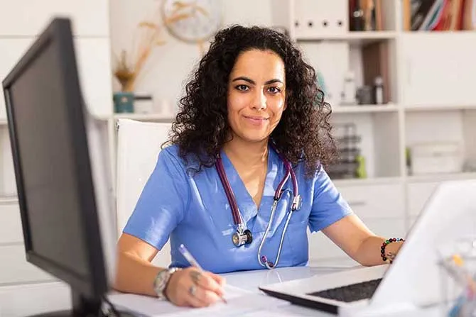 A woman in scrubs and a stethoscope works at a desk.