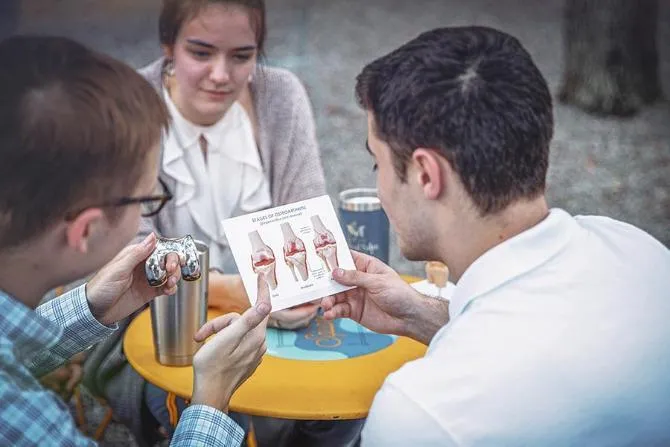 A young man holds a metal joint while looking at a card with text that says 'stages of osteoarthritis'