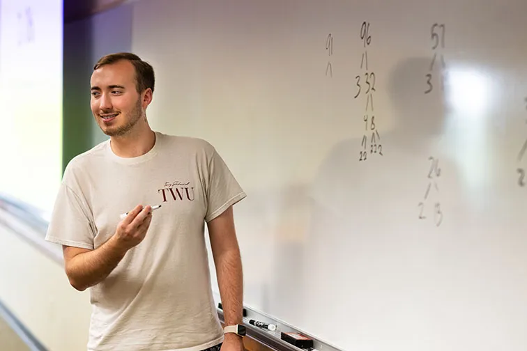 A math tutor instructs students in front of a whiteboard with math problems on it.