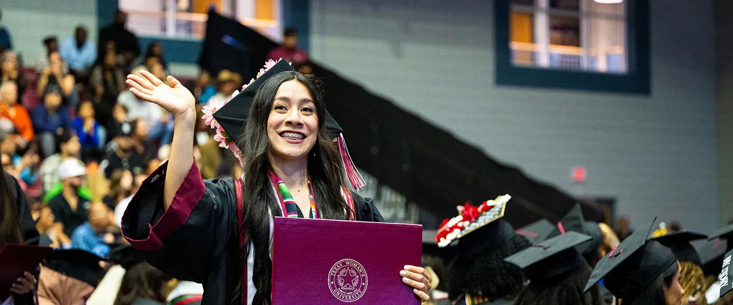 A woman in academic regalia waves to the crowd at her commencement ceremony.