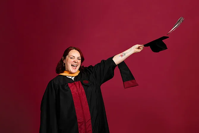 A portrait of a TWU student in graduate school academic regalia and holding a cap in the air.