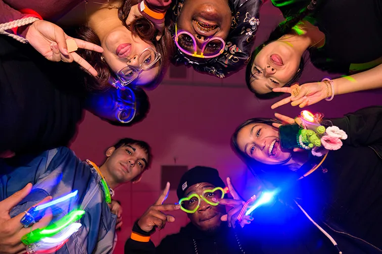 Students in a circle pose for a photo with blacklight glasses and accessories during an on campus event.