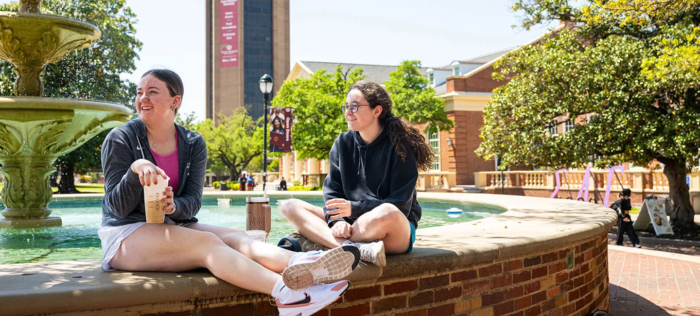 Two students hang out at the Denton campus fountain on a sunny day.