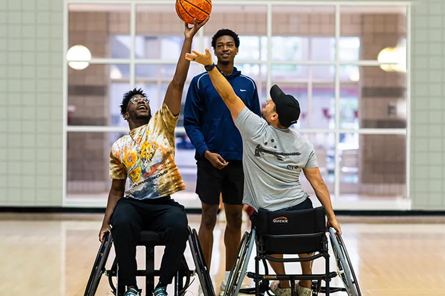 A TWU kinesiology student stands on a basketball court with two players in wheelchairs.