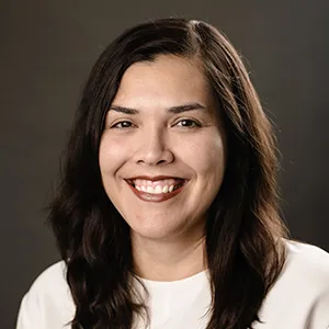 Headshot of Christine Adame. Women with dark hair, smiling. White top. Brown background.