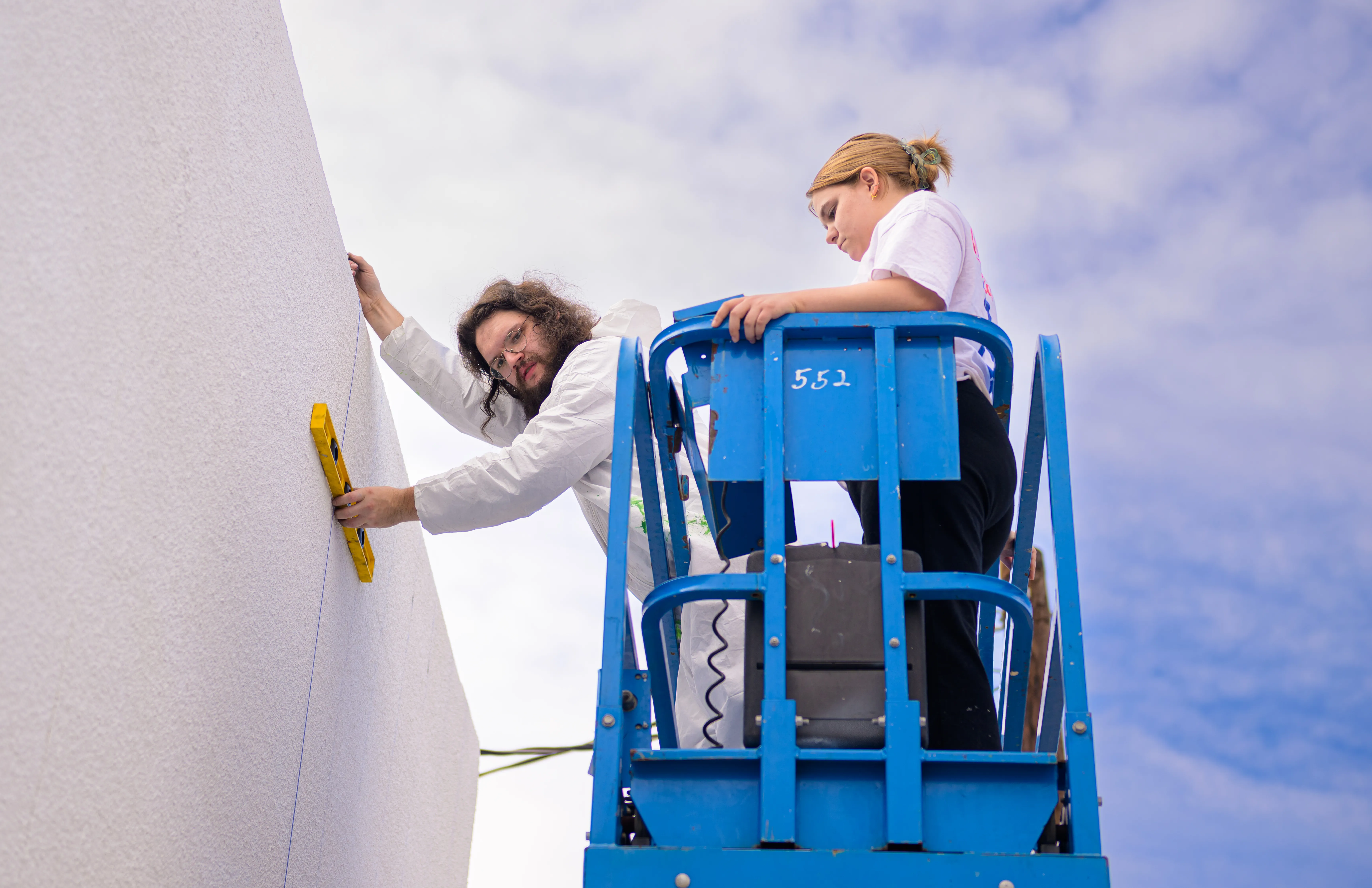 Students on scissor lift work on new mural, against blue sky