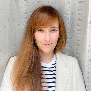Headshot of Meg Griffiths. Woman in white blazer and striped shirt. White curtain in the background.