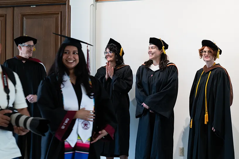 BFA in Graphic Design graduate, Citlalli Alaniz, walks through the TWU foyer in her commencement cap & cown. TWU faculty, Colby Parsons, Julie Libersat, Christine Adame, and Shelli Petersen cheer behind her.