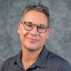 Headshot of Giovanni Valderas, 2023. Hispanic man in dark grey polo, wearing glasses, and curly hair