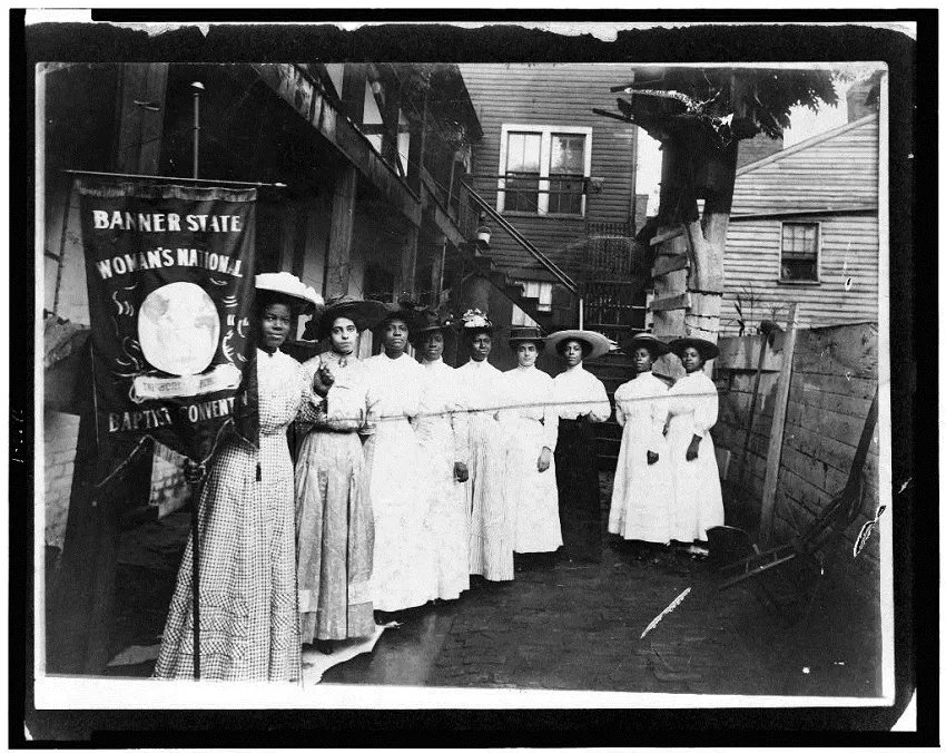 Nine African-American women posed, standing, full length, with Nannie Burroughs holding banner reading, 