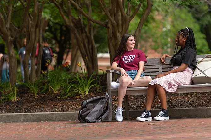 Two students talk on a bench outdoors on campus.