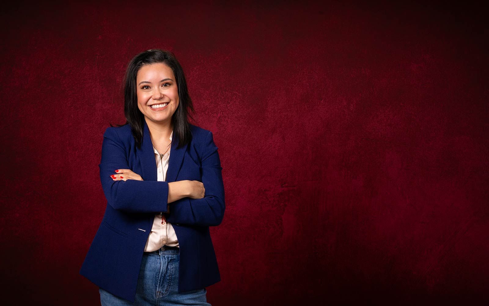 TWU student wearing jeans and a blazer smiles in front of a maroon backdrop