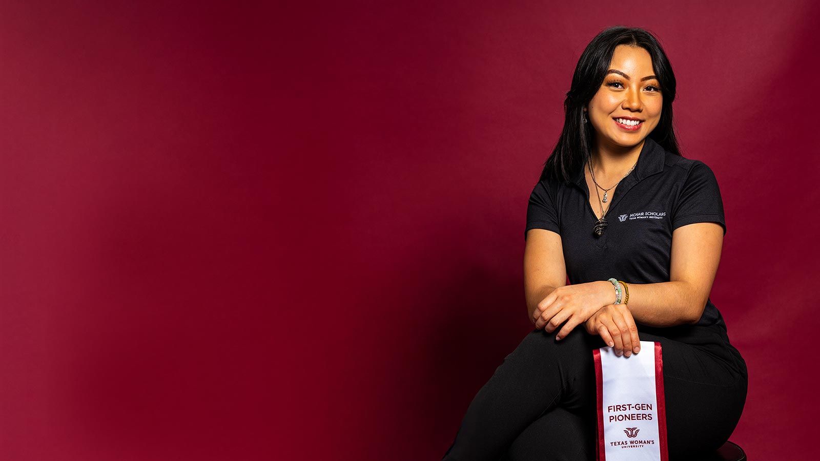 A TWU student dressed in black and holding up a white ribbon with the words 'First Generation' in maroon letters