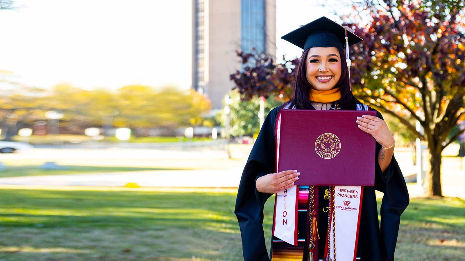 A TWU student in graduation regalia holding up a diploma