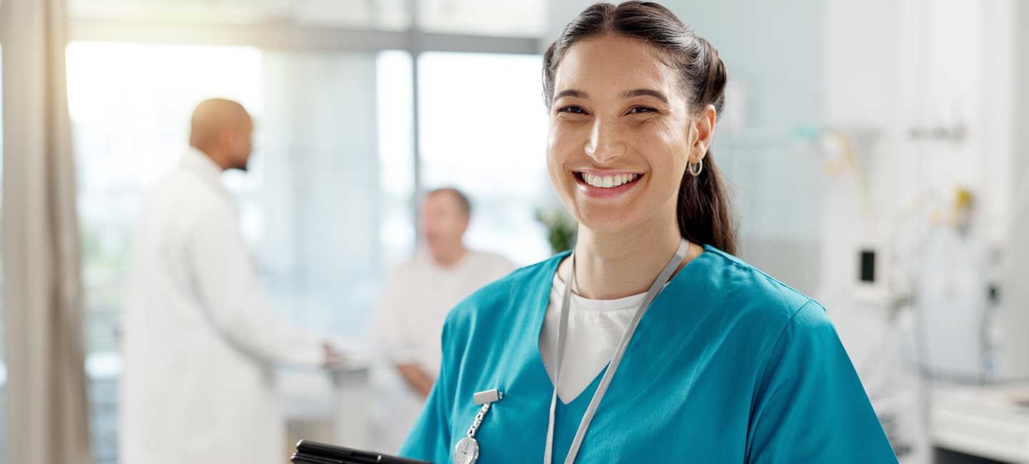 A woman smiles at the camera wearing scrubs and in a medical setting.