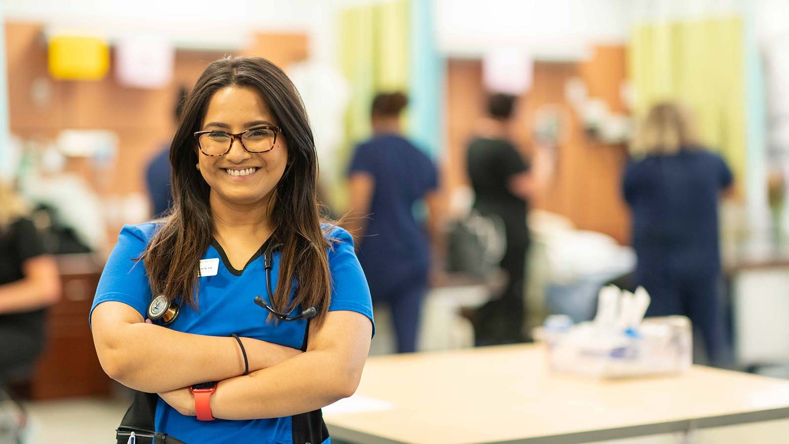 A young woman ins scrubs with a stethoscope around her neck smiles in a medical lab setting